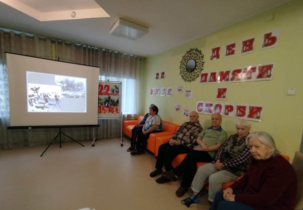 пожилые люди смотрят презентацию в зале, оформленном ко Дню памяти и скорби, расположившись на диванах