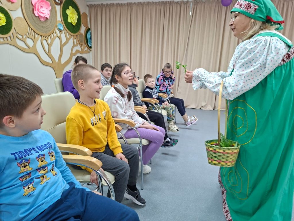 Специалист в русском народном костюме Травницы Агафьи держит в руках корзинку и показывает лекарственные травы группе детей