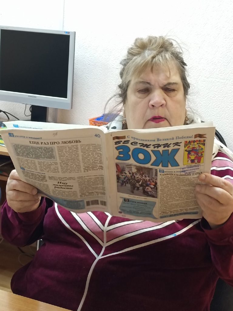 Женщина сидит на стуле за столом и читает журнал "Вестник ЗОЖ"