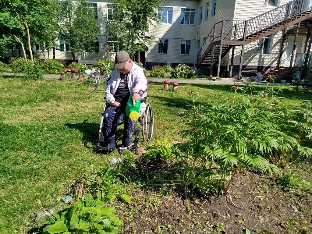 получатель услуг поливает из лейки большую клумбу с цветами