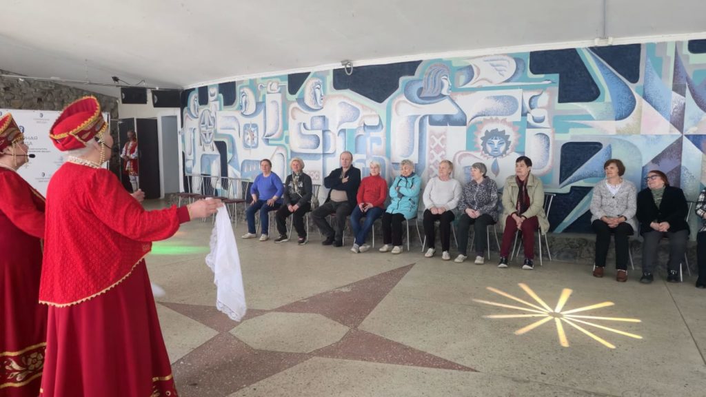 Мужчины и женщины , в количестве 10 человек слушают выступление выступление ансамбля русской песни Оленегорочка