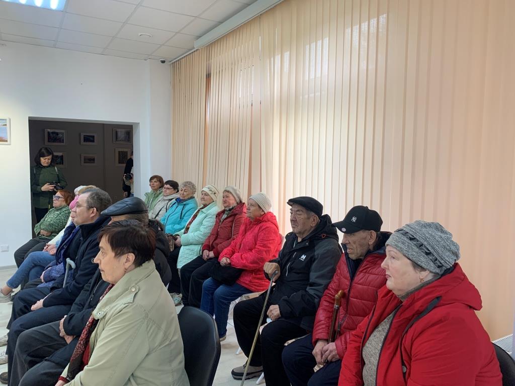 Мужчины и женщины, в количестве 15 человет сидят, слушают гида музейно-выстовочного зала У Оленьей горы